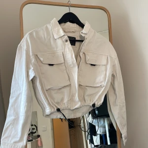 Vit croppad jeansjacka FB Sister - Snygg vit croppad jeansjacka från FB Sister med dragkedja framtill, stora bröstfickor och justerbar snörning i midjan. Jackan har klassisk krage och långa ärmar med knapp vid ärmslut. Perfekt för dig som gillar streetstyle och vill sticka ut.