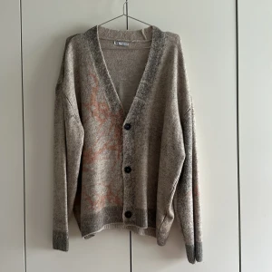 Beige kofta med tryck från Zara - Säljer en beige kofta från Zara med unikt tryck i grönt och rostbrunt på ryggen och framsidan. Koftan har v-ringning, knappar framtill och långa ärmar. Materialet är mjukt och stickat, perfekt för lager-på-lager under kyliga dagar.