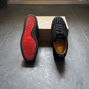 Svart christian loubitins - Svarta sneakers från Christian Louboutin i mocka med ikonisk röd sula och svarta nitar på tån. Skorna har snörning och rund tå, samt exklusiv känsla med märkets signaturdetaljer. Perfekta för dig som vill sticka ut med edgy och lyxig stil.
