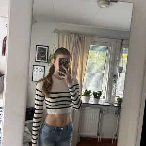 Randig croppad stickad tröja - Säljer en croppad, ribbstickad tröja med svarta ränder på beige botten. Tröjan har lång ärm, rund halsringning och sitter tight. Perfekt för dig som gillar en trendig och clean look.