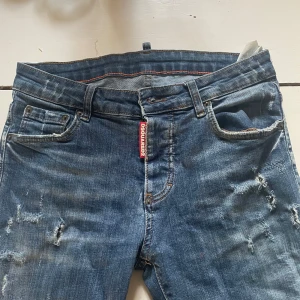 Dsquared2 blå slitna jeans - Säljer ett par blå jeans från Dsquared2 med snygga slitningar och distressed detaljer. Klassisk femficksmodell med röd Dsquared2-tag vid gylfen. Jeansen har normal passform och är tillverkade i bomull för en skön känsla.