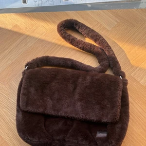 Brun fluffig axelväska i fuskpäls - Mysig brun axelväska i mjuk fuskpäls med bred axelrem. Väskan har ett stort huvudfack och en innerficka med dragkedja. Perfekt för dig som vill ha en trendig och gosig accessoar till din outfit.