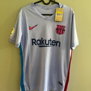 FC Barcelona bortatröja Nike ljuslila - FC Barcelona bortatröja från Nike i ljuslila polyester med mörkblå Rakuten-tryck och klubbmärke i rosa/rött. Kortärmad modell med V-ringning, La Liga-märke på ärmen och färgdetaljer i blått och rött längs sidorna. Perfekt för dig som älskar Barca!