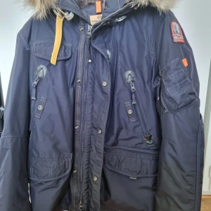 Parajumpers right handed parkas med päls - Mörkblå parkas från Parajumpers med snygg päls på huvan, flera praktiska fickor och ikonisk PJS-patch på ärmen. Jackan har quiltat foder, dragkedja och knappar framtill samt cool gul detalj vid kragen. Perfekt för kalla dagar och riktigt street.