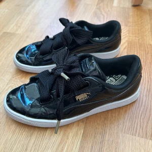 Svarta Puma Basket sneakers lack - Snygga svarta Puma Basket sneakers i glansigt lackmaterial med breda svarta snören och vit sula. Guldiga Puma-loggor på sidan och hälen. Skorna har rund tå och klassisk sneakerform, perfekt för dig som gillar statement-detaljer.