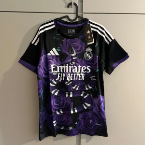 Real Madrid x Adidas svart/lila fotbollströja - Snygg Real Madrid fotbollströja från Adidas med svart bas, lila rosor och en stor ormmönstrad print på framsidan. Klassiska vita Adidas-ränder på axlarna och klubbmärke på bröstet. Tillverkad i lätt och ventilerande material.