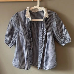 Randig skjorta Holly & Whyte Lindex - Snygg blåvit randig skjorta från Holly & Whyte by Lindex med kort puffärm och vit volangkrage. Skjortan har knappar framtill, perfekt för en fräsch och stilren look.