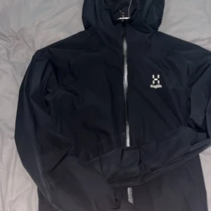 Haglöfs jacka  - Pris kan diskuteras, skicka dm för video på jackan. Svart regnjacka från Haglöfs med minimalistisk design och broderad logga på bröstet. Jackan har huva, dragkedja framtill och två sidofickor. Perfekt för blöta dagar och enkel att slänga på sig när vädret sviker. Storlek ”M”