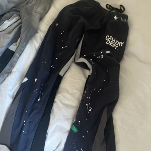 Svarta mjukisbyxor Gallery Dept. - Svarta mjukisbyxor från Gallery Dept. med vit logga på låret och coola färgstänk i vitt och grönt. Byxorna har elastisk midja med snörning och är tillverkade i mjukt material för maximal komfort. Perfekta för en avslappnad och trendig look.