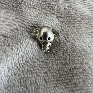 Silverfärgad Dobby-berlock - Söt berlock i form av Dobby från Harry Potter, gjord i silverfärgad metall. Perfekt att trä på ett armband eller halsband för en personlig touch. Detaljerad design med stora öron och utstansade ögon, hål genom huvudet för enkel montering. Funkar till Pandora armband.