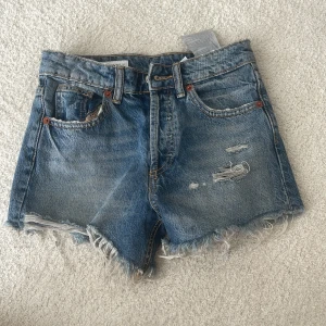 Blå jeansshorts med fransig kant - Snygga blå jeansshorts med fransig kant nertill. Shortsen har slitna detaljer framtill och klassiska fem fickor. Perfekta för sommaren och ger en avslappnad vibe till din outfit.