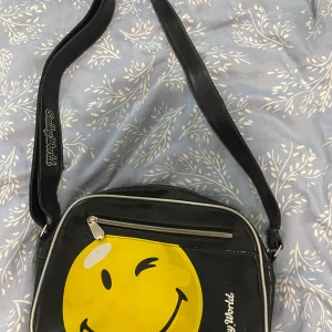 Svart axelväska SmileyWorld retro - Cool svart axelväska från SmileyWorld med stort gult smileytryck på framsidan. Väskan har bred axelrem med logga, vita kanter och dragkedja både fram och upptill. Tillverkad i syntetmaterial med retrovibbar och plats för allt du behöver.