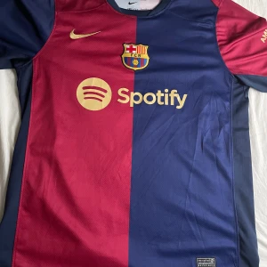 FC Barcelona Nike fotbollströja blå/röd - Snygg FC Barcelona matchtröja från Nike med klassisk halva-halva design i mörkblått och vinrött. Tröjan har korta ärmar, klubbmärke på bröstet och guldfärgade detaljer som Nike-logga och Spotify-tryck. Tillverkad i lätt och ventilerande material perfekt för fotboll.