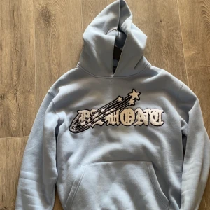 Ljusblå hoodie från Demont med tryck - Säljer en ljusblå hoodie från Demont i storlek S. Tröjan har ett stort broderat tryck med stjärnor och texten 'Demont' på bröstet. Använd Max 3-4 gånger är som helt ny dom finns inte och köpa längre ,storlek s men sitter som M