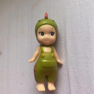 Sonny angel  - Bilderna visar en leksaksfigur utklädd i en grön dinosauriedräkt med röda och gula detaljer. Ingen bok syns på bilderna.