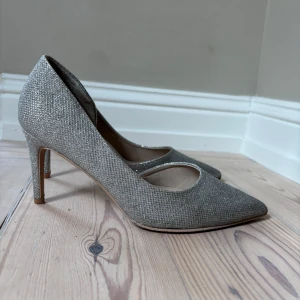 Glittriga silverfärgade pumps (storlek 38) - Snygga pumps i glittrigt silver med spetsig tå och hög klack. Skorna är bara använda en gång på bal men är i ett jättefint skick. Är perfekta om man vill klä upp en basic outfit.