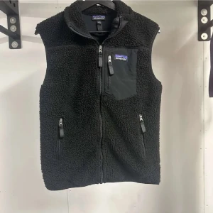 Svart fleeceväst från Patagonia S - Svart fleeceväst från Patagonia med hög krage, tre dragkedjeförsedda fickor och mjuk teddystruktur. Västen har en bröstficka med logga och är perfekt för lager-på-lager. Snygg och funktionell för kyliga dagar.