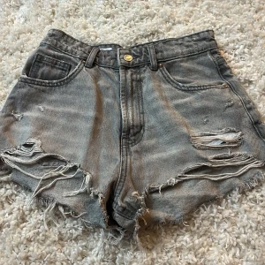 Gråa ripped jeansshorts från Zara - Snygga gråa jeansshorts från Zara med hög midja och råa, fransiga kanter. Shortsen har slitna detaljer och flera revor framtill för en cool, avslappnad vibe. Klassisk femficksmodell och dragkedja med knapp. Perfekta för sommardagar när du vill ha en edgy look.