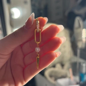 Guldigt örhänge med pärla och strass - Snyggt örhänge i guld med glittrande strass upptill, en avlång länk, en vit pärla i mitten och en spetsig guldig detalj längst ner. Perfekt statement-piece för dig som gillar unika smycken.