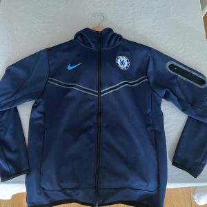 Chelsea Nike Tech - Snygg marinblå vindjacka från Nike med Chelsea FC-logga på bröstet. Jackan har huva, hel dragkedja och diskreta grå detaljer över bröstet samt en ficka med dragkedja på ärmen. Perfekt för dig som älskar fotboll och vill representera ditt lag.