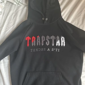 Svart Trapstar hoodie med brodyr - Svart hoodie från Trapstar med stor broderad logga i rött, grått och vitt på bröstet. Klassisk huva med snörhål och känguruficka framtill. Mjuk insida och relaxed fit, perfekt hoodie för alla årstider.
