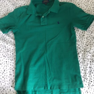 Grön pikétröja från Ralph Lauren - Snygg grön pikétröja från Polo Ralph Lauren i klassisk modell med krage och två knappar. Tröjan har korta ärmar och den ikoniska broderade loggan på bröstet. Tillverkad i mjuk bomull som är skön mot huden. Litet hål vid kragen som tecks av kragen och kan sys 