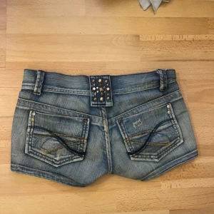 Lågmidjade jeansshorts med nitar - Säljer dessa assnygga lågmidjade jeansshorts i klassisk blå denim med slitningar och coola detaljer då de tyvärr har blivit för små för mig. Baksidan har dekorativa sömmar och nitar på linningen. Shortsen har fyra fickor, bälteshällor och dragkedja framtill. Perfekta för en avslappnad och trendig look.