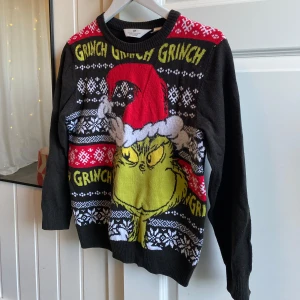 Jultröja från H&M - Svart jultröja från H&M med stort Grinch-motiv och texten 'Grinch' i gult. Tröjan har röda och vita detaljer, snöflingor och julmönster. Långärmad och ribbad vid hals, ärmslut och nederkant. Perfekt för dig som vill sticka ut på julfirandet!