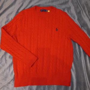 Röd kabelstickad tröja Polo Ralph Lauren - Säljer en röd kabelstickad tröja från Polo Ralph Lauren. Tröjan har rund hals, långa ärmar och klassisk broderad logga i blått på bröstet. Ribbstickade muddar vid ärmslut och nederkant. Perfekt för dig som gillar stilren och tidlös design.