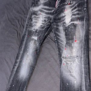 Dsquared2 svarta skinny jeans med färgstänk - Säljer ett par svarta skinny jeans från Dsquared2 med slitningar, trasiga detaljer och coola färgstänk i rött och vitt. Jeansen har knappgylf, klassisk femficksmodell och är tillverkade i bomull. Perfekt för dig som gillar en edgy streetstil.