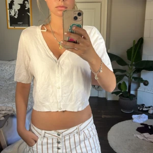 Vit croppad kofta från STOCKH LM - Säljer en vit croppad blus från STOCKH LM med korta ärmar och v-ringning. Toppen har knappar framtill och är tillverkad i ett mjukt bomullsmaterial. Perfekt att styla med jeans eller kjol