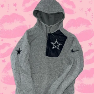 Grå Dallas Cowboys hoodie från Nike - Snygg grå hoodie från Nike x NFL Dallas Cowboys med stor stjärnlogga på bröstet och svart detaljpanel. Tröjan har huva med snörning, lång ärm och Nike-logga på ärmen. Perfekt för dig som gillar sportig stil och vill representera Cowboys.