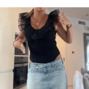Volang body - Supersnygg svart linnetopp med transparenta volangaxlar och djup v-ringning. Toppen är tight och figurnära, perfekt att styla med jeans eller kjol för en trendig look.