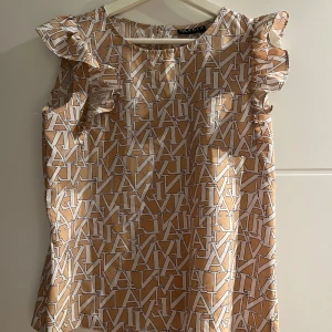 Beige blus med volang från Soho Fashion - Säljer en beige blus från Soho Fashion med coolt grafiskt mönster i vitt. Blusen är ärmlös med volangdetaljer vid axlarna och har rund halsringning. Materialet är glansigt och känns lyxigt, perfekt för dig som gillar statement-plagg.