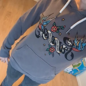 Grå Superdry Hoodie - Grå märkeshoodie från Superdry med snyggt glittrigt och coolt motiv fram. Tröjan har huva med vita snören. Perfekt för dig som gillar streetstyle och y2k. Säljer för ett lite högre pris eftersom den är unik och köptes dyrt. 