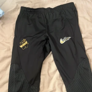 Svarta AIK Nike träningsbyxor M - Svarta träningsbyxor från Nike med AIK-logga i guld och vitt på ena benet och Nike-logga i guld på andra. Byxorna har elastisk midja, paneler med mönster vid knäna och är gjorda i lätt polyester med Dri-FIT-teknik. Perfekta för fotbollsträning eller chill.