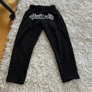 Svarta mjukisbyxor från Hoodrich XS - Svarta mjukisbyxor från Hoodrich i storlek XS. Byxorna har elastisk midja med snörning och en stor vit Hoodrich-logga framtill. Sköna och stilrena, perfekta för chill eller streetwear. Tillverkade i mjukt material för maximal komfort.