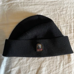 Svart ribbstickad mössa Parajumpers - Svart ribbstickad mössa från Parajumpers med uppvikt kant och klassisk PJS-logga framtill. Snygg och enkel design som passar perfekt till kalla dagar. Tillverkad i mjukt material som håller dig varm och bekväm. Detta är den tunnare modellen av parajumper mössorna.