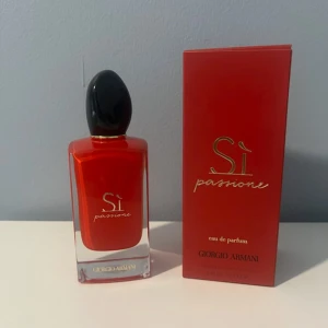 Si Passione Parfym Giorgio Armani - En elegant och passionerad parfym i en vacker röd glasflaska med svart kork. Flaskan har en modern, fyrkantig form med tydlig märkning av parfymens namn och märke. Förpackningen är i matchande röd färg med gulddetaljer och tydlig text om innehåll och ingredienser. Perfekt för den som vill ha en doft som utstrålar självsäkerhet och stil.