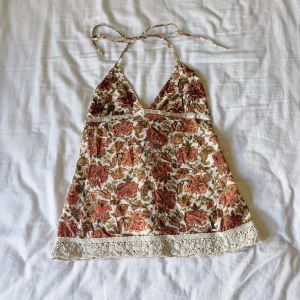 Blommig halterneck linnetopp med spets - Supersöt linnetopp med blommigt mönster i beige, rött och brunt. Halterneck med knyt i nacken och ryggen. Dekorativ spetskant nertill och pärldetaljer vid brösten. Perfekt för sommarens varma dagar. Storlek 38 på lappen men mer som en 36. 