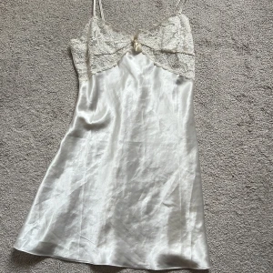 Vit satin slipklänning med spets - Superfin kort vit slipklänning i satin med tunna axelband och spetsdetaljer upptill. Söt liten rosett framtill och blommig spets ger en romantisk vibe. Perfekt för sommarens varma dagar eller som layering-piece. Det är storlek 38/40 men passar även dom som har xs/s😊