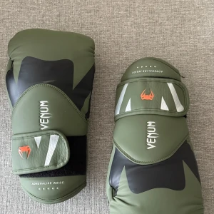 Venum boxningshandskar Grön 12oz - Säljer ett par boxningshandskar från Venum i olivgrönt och svart med vita detaljer och orange logga. Handskarna har coola grafiska mönster och bred kardborrestängning för extra stöd. Perfekta för träning och sparring.  OBS Vänster handske är sönder vid tummen… men den går fortfarande att använda och stör ej under användning. 10 månader gammal 