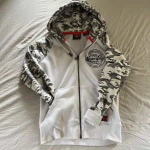 Superdry hoodie med camoärmar, M - Vit hoodie från Superdry med grå och vita camouflageärmar och huva. Stor logga på bröstet och dragkedja framtill. Hoodien har kängurufickor och coola orangea detaljer på insidan av huvan. Perfekt för dig som gillar streetstyle.
