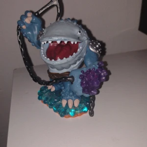Skylander figur Haj  - Bilderna visar en leksaksfigur som liknar ett blått monster med stora tänder, kedjor och lila kristaller. Figuren står på en genomskinlig blå och orange bas. Ingen bok syns på bilderna.