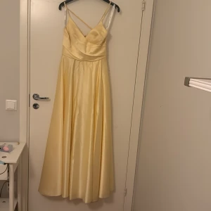 Ljusgul långklänning med slits - Elegant ljusgul långklänning i satinliknande material med smala axelband, v-ringning och omlottliv. Klänningen har öppen rygg med snygg snörning och hög slits framtill för en extra touch. Perfekt för bal eller bröllop. Skulle säga att den även passar en i storlek 36 eftersom man spänner banden för rätt storlek. Har ändats används en gång, prislapp finns även kvar. Pris kan diskuteras 😊