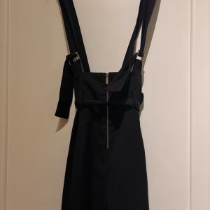 Formula Joven Svart overall med rosett - Supersnygg svart lång overall från Formula Joven med breda axelband, dragkedja framtill och en stor rosett i midjan. Overall har en stilren look och är perfekt för dig som gillar minimalistisk stil med en twist.