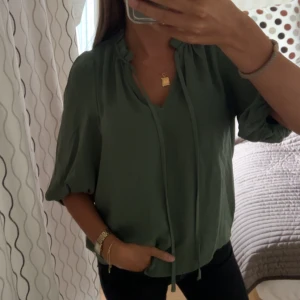 Grön blus med knyt och volang - Snygg grön blus från Vero Moda med volangdetalj vid halsen och knytband framtill. Blusen har trekvartsärmar med resår vid ärmslut och en avslappnad passform. Perfekt för dig som gillar en chill men ändå stilsäker look.