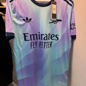 Arsenal x Adidas lila-blå fotbollströja - Snygg Arsenal fotbollströja från Adidas med vågigt mönster i lila och ljusblått. Tröjan har mörkblå detaljer vid krage och ärmslut, samt tryck med Emirates och klubbens logga på bröstet. Tillverkad i lätt och ventilerande material, perfekt för match eller träning.