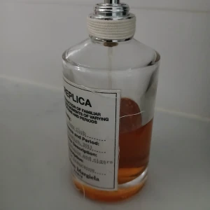 Maison Margiela Replica parfym - Maison Margiela Replica parfym i genomskinlig glasflaska med vit etikett och silverfärgad spraytopp. Parfymen har en varm bärnstensfärgad vätska och klassisk minimalistisk design. Perfekt för dig som gillar stilrena och ikoniska dofter.
