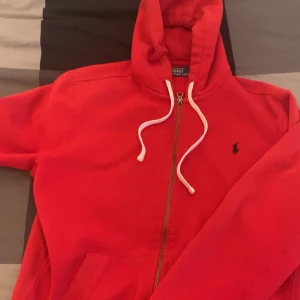 Röd hoodie från Polo Ralph Lauren - Riktigt snygg röd hoodie från Polo Ralph Lauren med dragkedja och vit snörning i huvan. Klassisk logga broderad på bröstet. Perfekt för dig som gillar streetstyle och vill ha något bekvämt och stilrent.